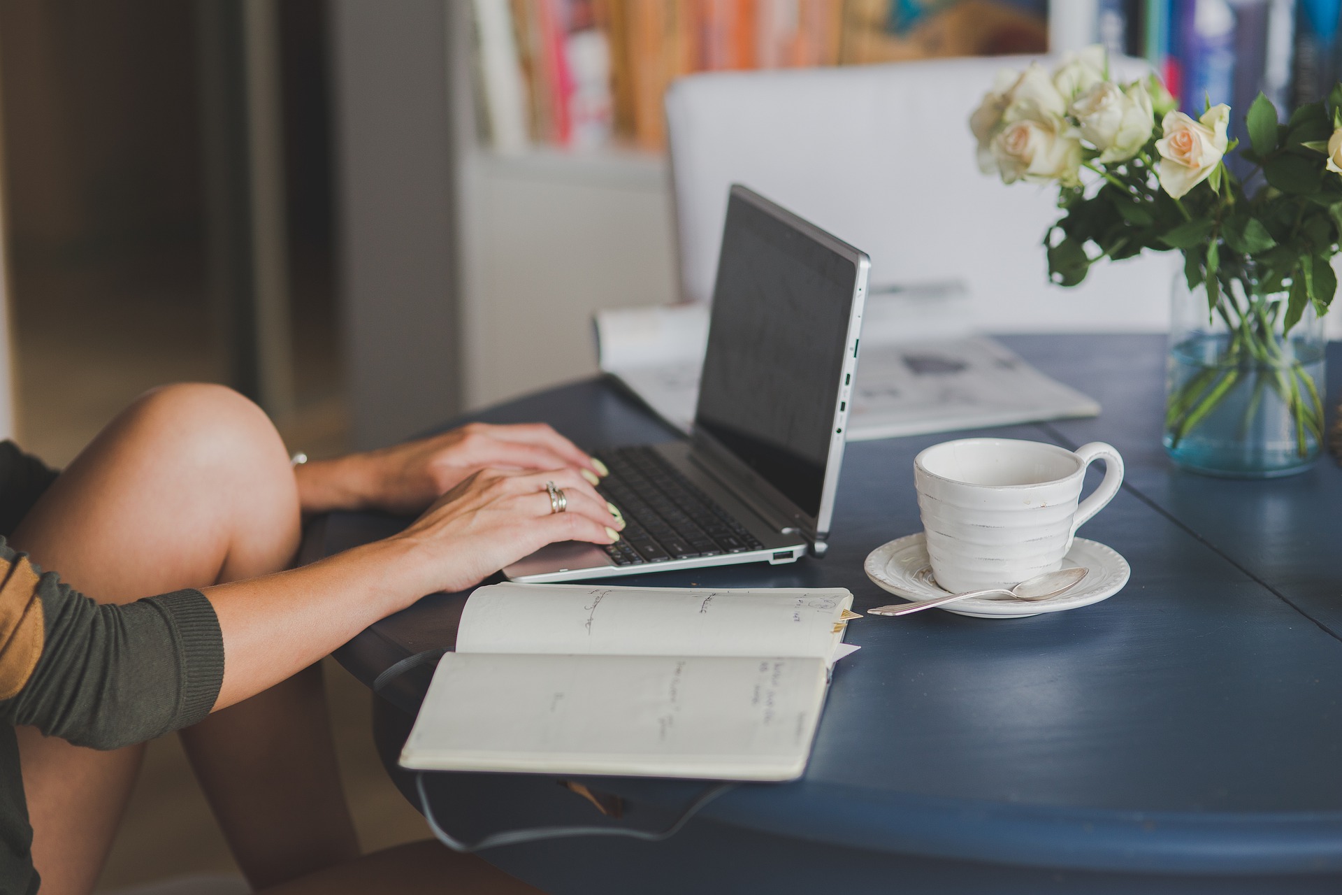 Woman sitting typing her SEO business blog on a laptop with a cup of tea and a notebook open on the table.